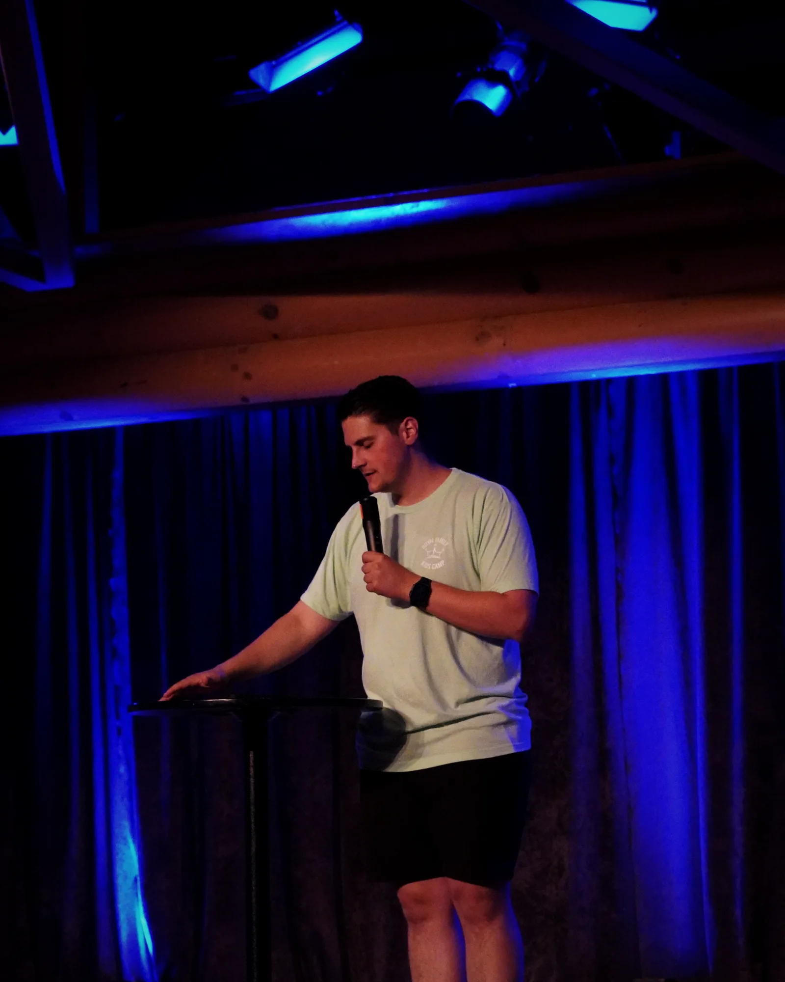 Carlo Orlando at a small podium on a camp stage lit in blue, microphone in hand
