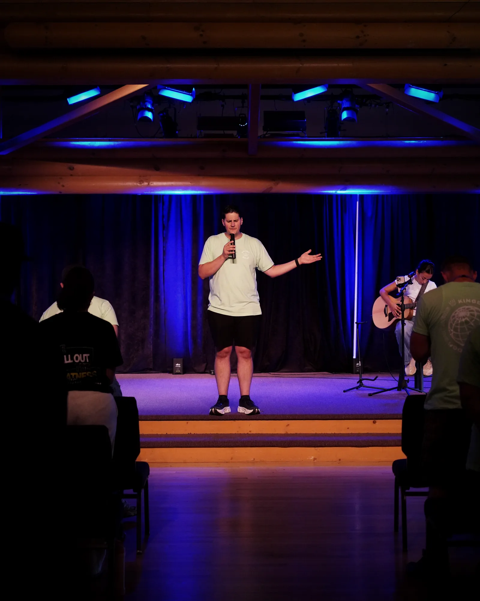 Carlo Orlando speaking on a camp stage under log beams and blue light, a guitarist beside him and campers in the foreground