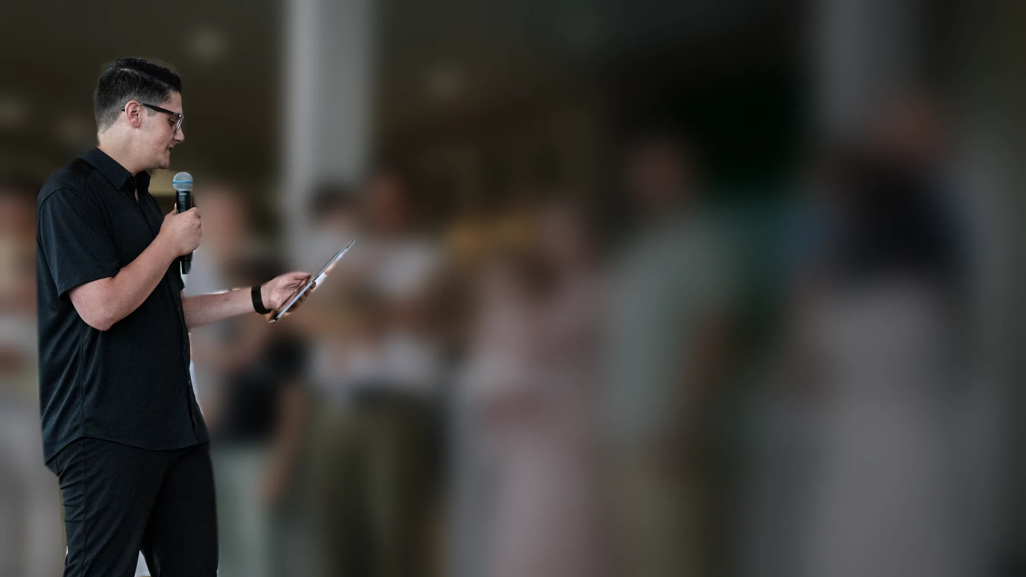 Carlo Orlando, microphone in hand, reading from a tablet during a family dedication; the families beside him are softly out of focus
