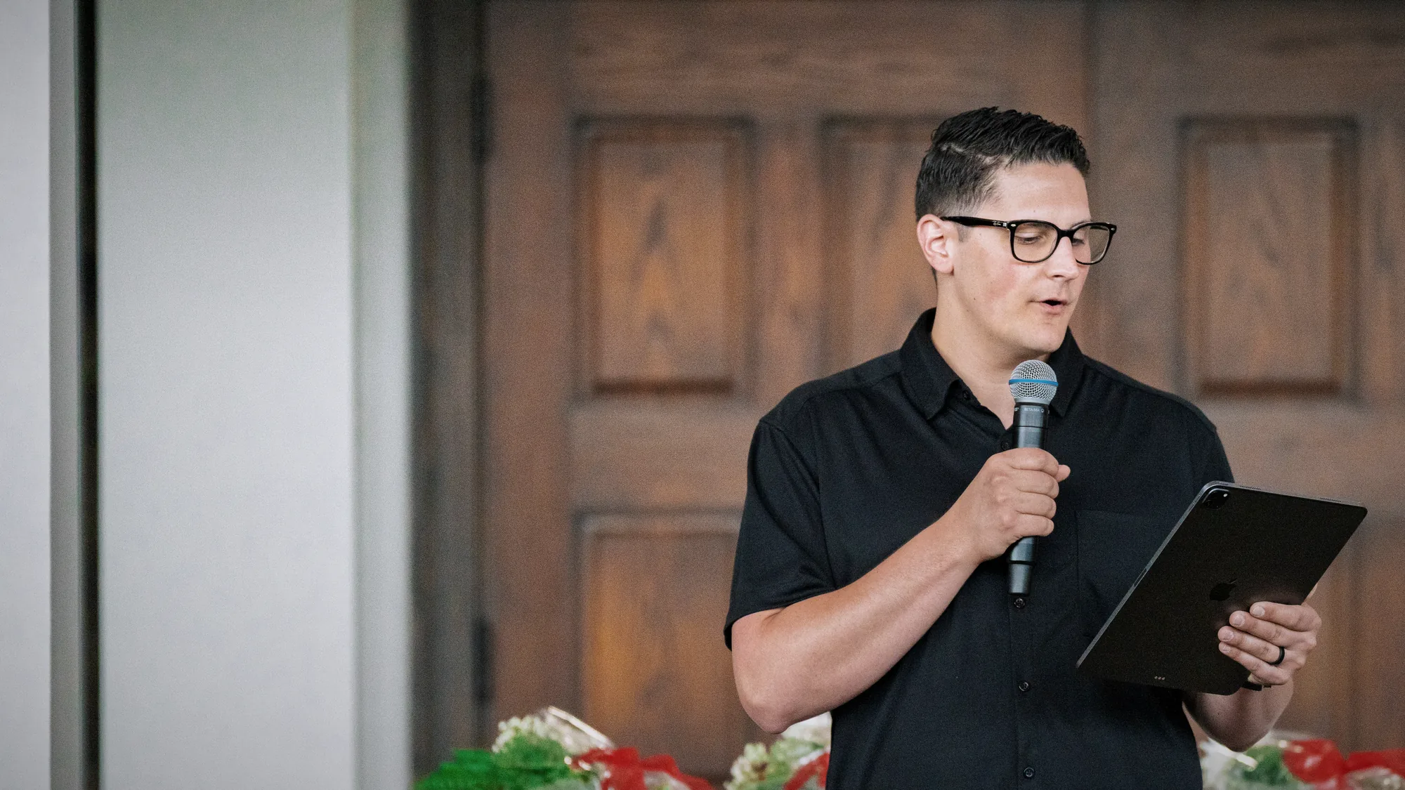 Carlo Orlando in glasses reading from a tablet into a microphone in front of tall wooden doors
