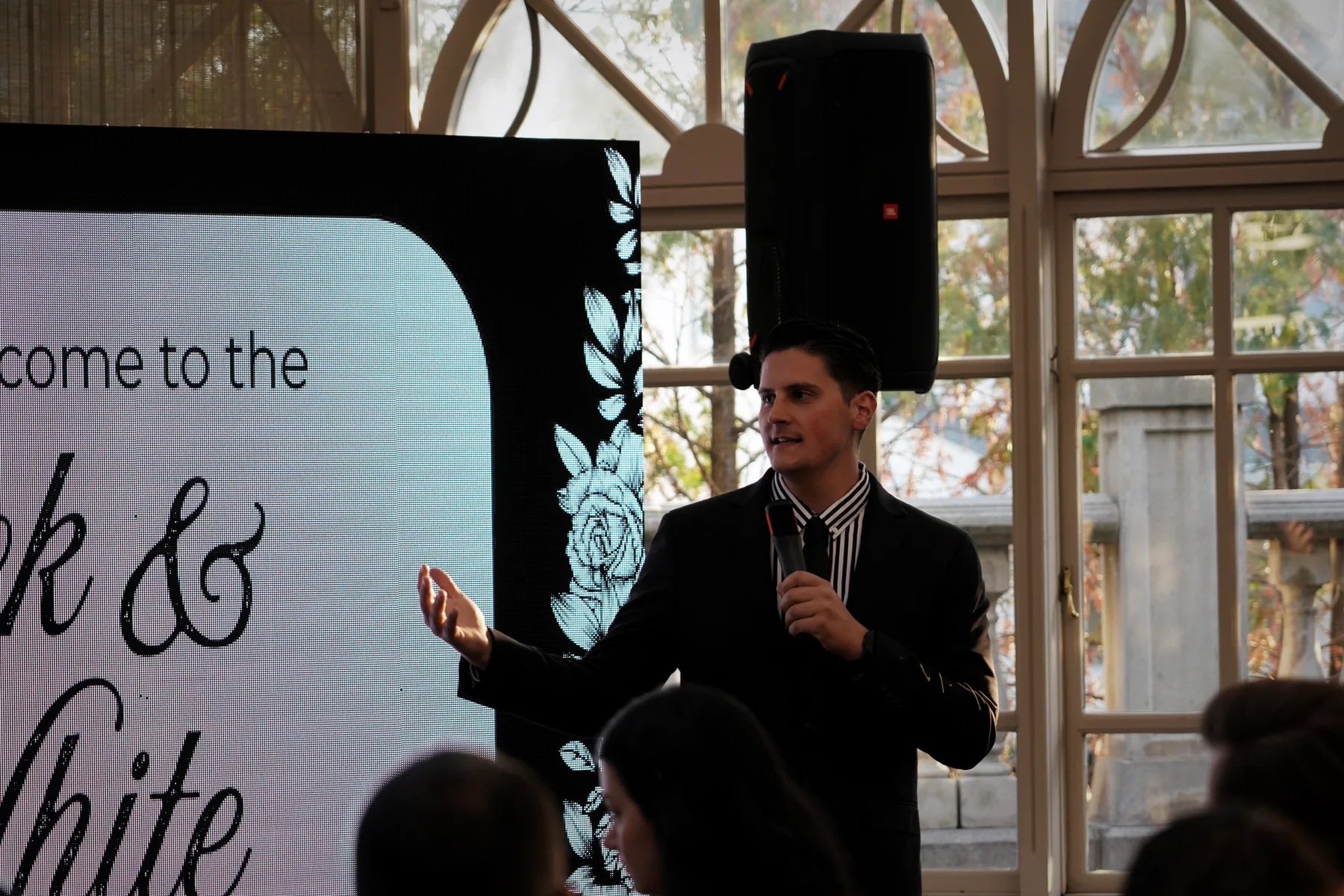 Carlo Orlando in a suit, microphone in hand, gesturing to a screen that reads Black & White, guests at their tables in the foreground
