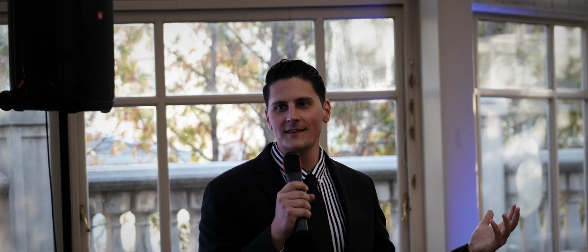 Carlo Orlando in a suit, smiling, one hand open toward the room as he speaks at the Black & White gala