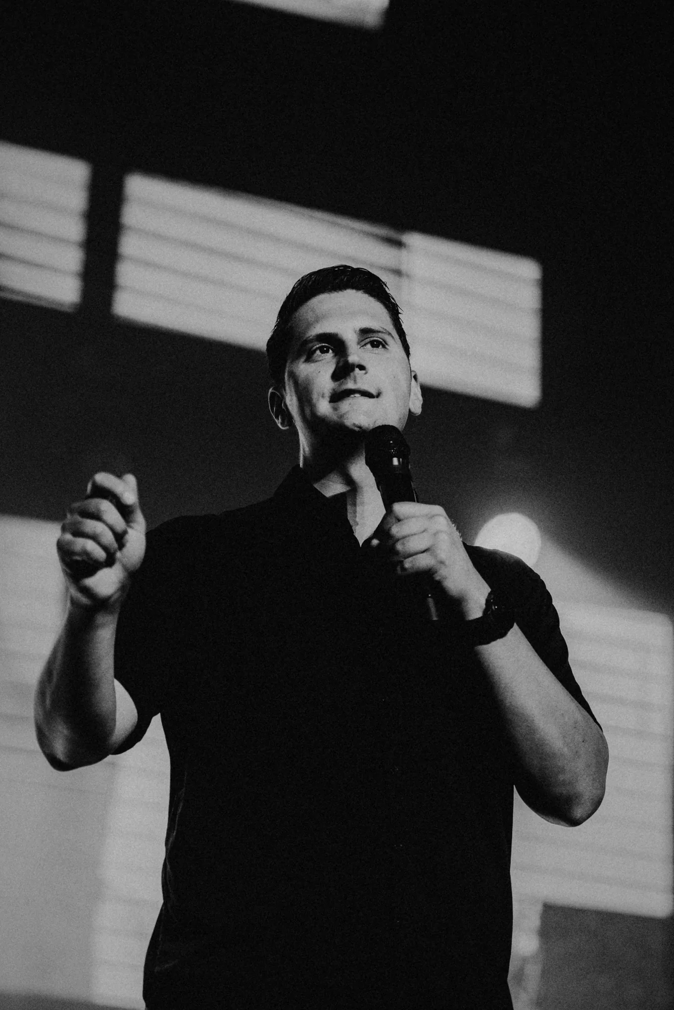 Black-and-white photograph of Carlo Orlando speaking into a microphone, one hand raised, in hard window light