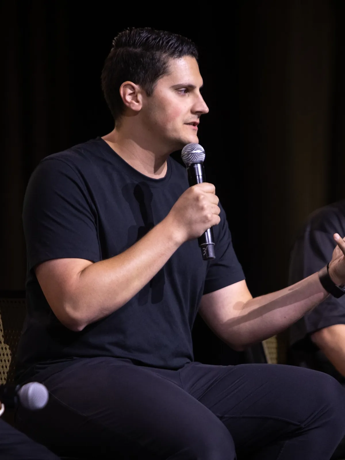 Carlo Orlando seated on a leadership panel, microphone in hand, turned to listen