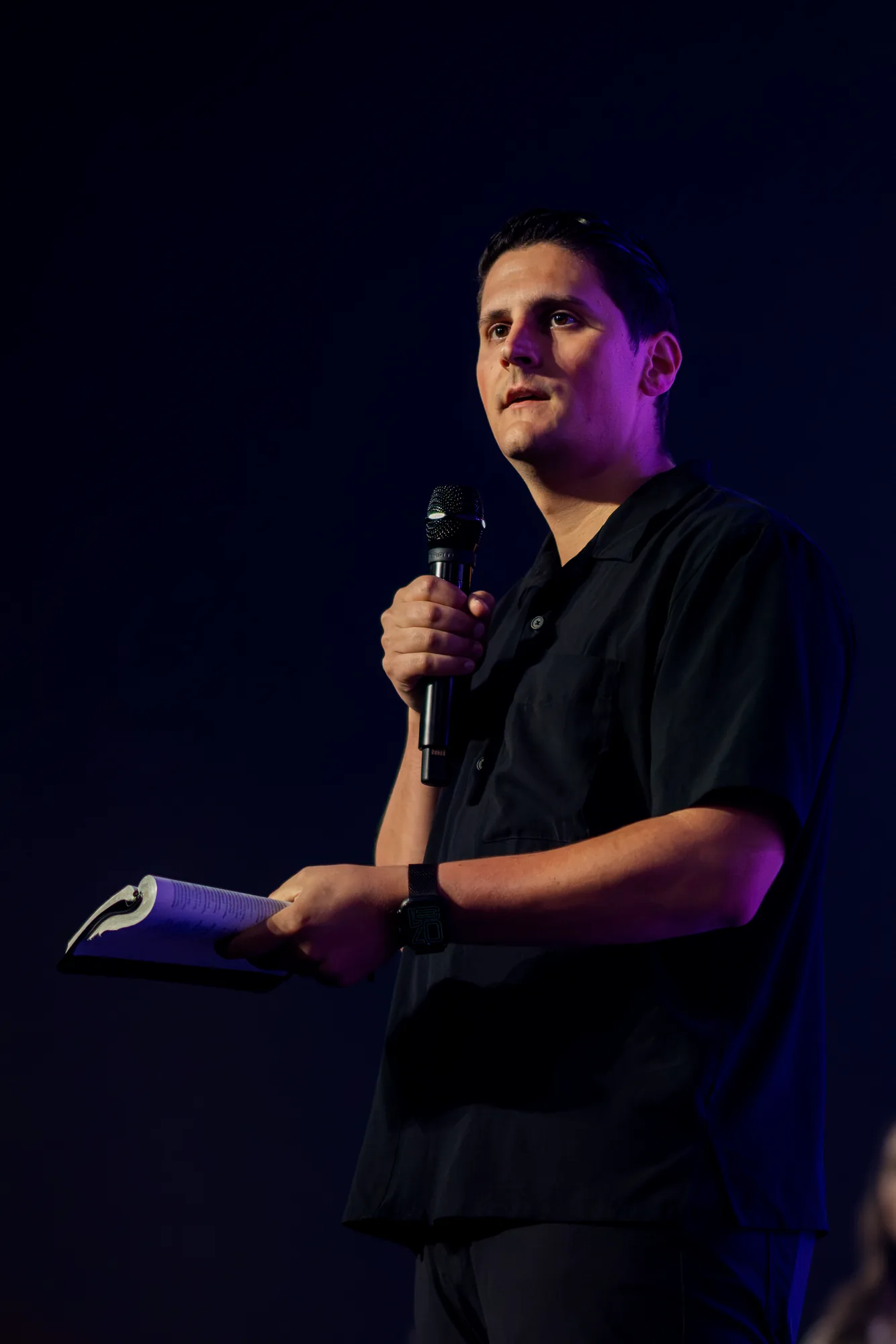 Carlo Orlando teaching on a dark stage, microphone raised and an open Bible in his hand