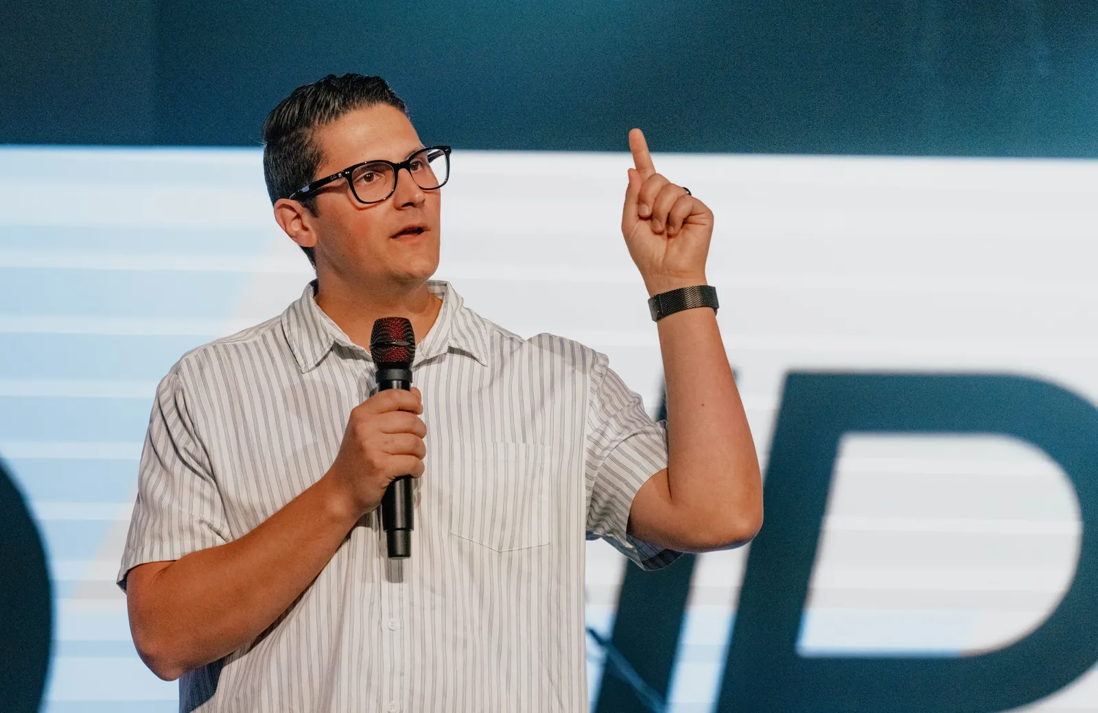 Carlo Orlando in glasses teaching on stage, microphone in one hand and the other pointing up